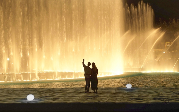 Couple watching Dubai Fountain show from boardwalk near Burj Khalifa at night.