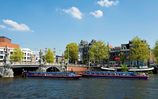 Canal cruise boats on Amsterdam canal with historic buildings and bridge in view.