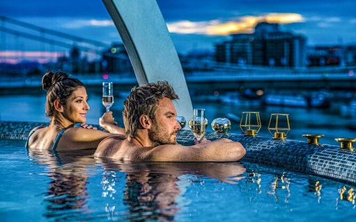 Couple enjoying Széchenyi Spa pool with city view, holding champagne glasses.