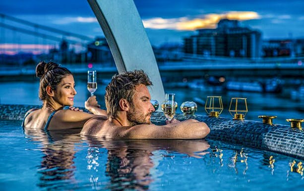 Couple enjoying Széchenyi Spa pool with city view, holding champagne glasses.