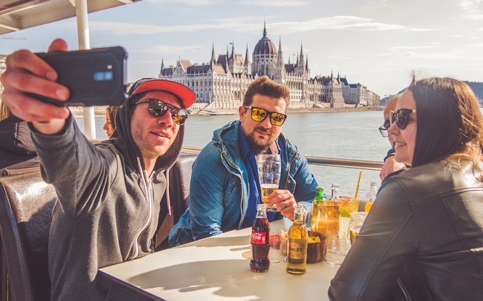 Group enjoying drinks on a Danube River cruise with Budapest Parliament in the background.