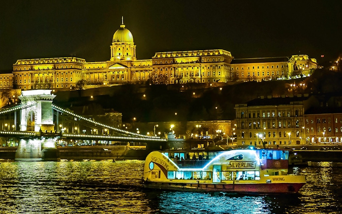 Danube River cruise boat passing by Budapest's illuminated Buda Castle at night.