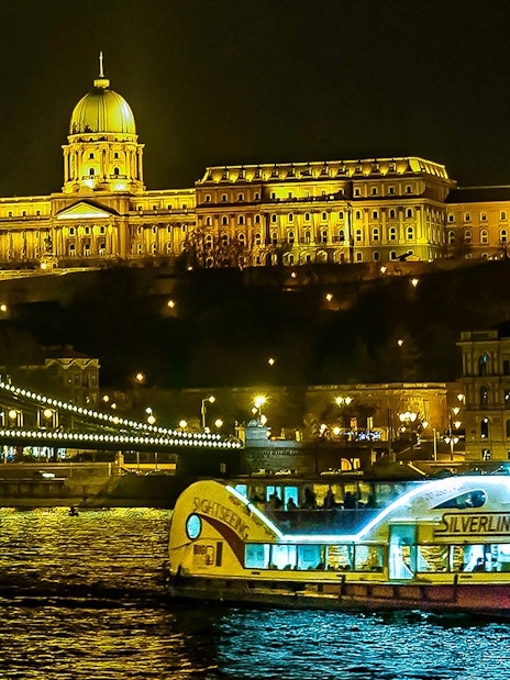 Danube River cruise boat passing by Budapest's illuminated Buda Castle at night.