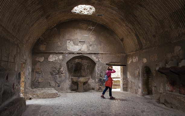 Visitor exploring ancient room in Herculaneum, Italy, with arched ceiling and stone basin.