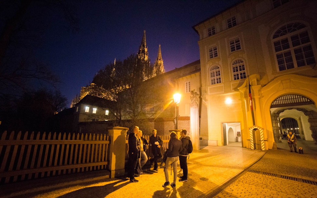 Group of people on a night tour near Prague Castle, illuminated spires in background.