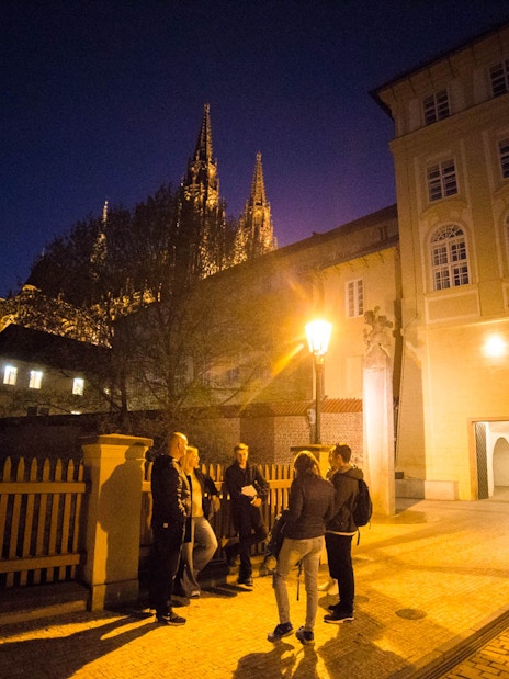 Group of people on a night tour near Prague Castle, illuminated spires in background.