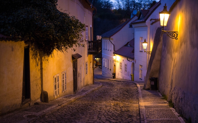 Narrow cobblestone street in Prague Castle area at dusk, illuminated by street lamps.