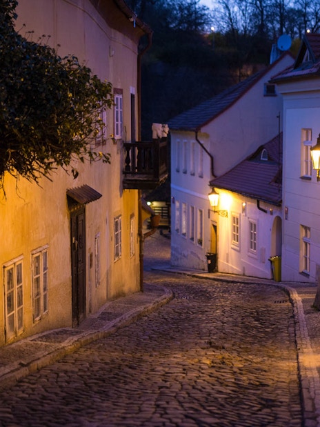 Narrow cobblestone street in Prague Castle area at dusk, illuminated by street lamps.