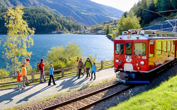Bernina Express train passing by a lake in the Swiss Alps with tourists walking nearby.