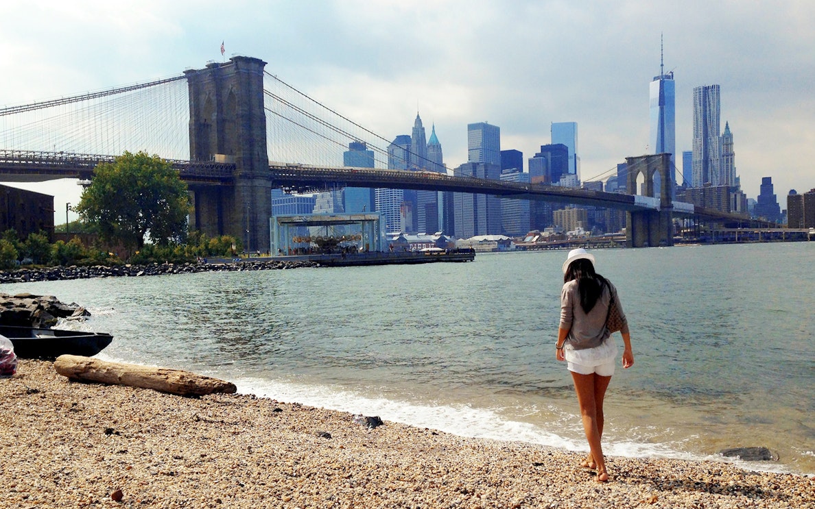 Brooklyn Bridge view from a pebbled beach, New York City skyline in the background.