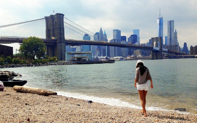Brooklyn Bridge view from a pebbled beach, New York City skyline in the background.