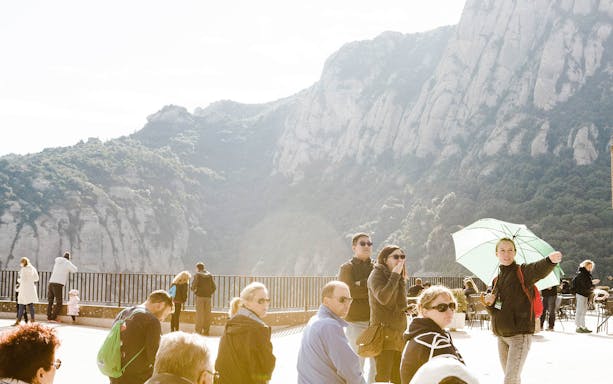 Tourists on Montserrat viewing platform with guide pointing, mountains in background.