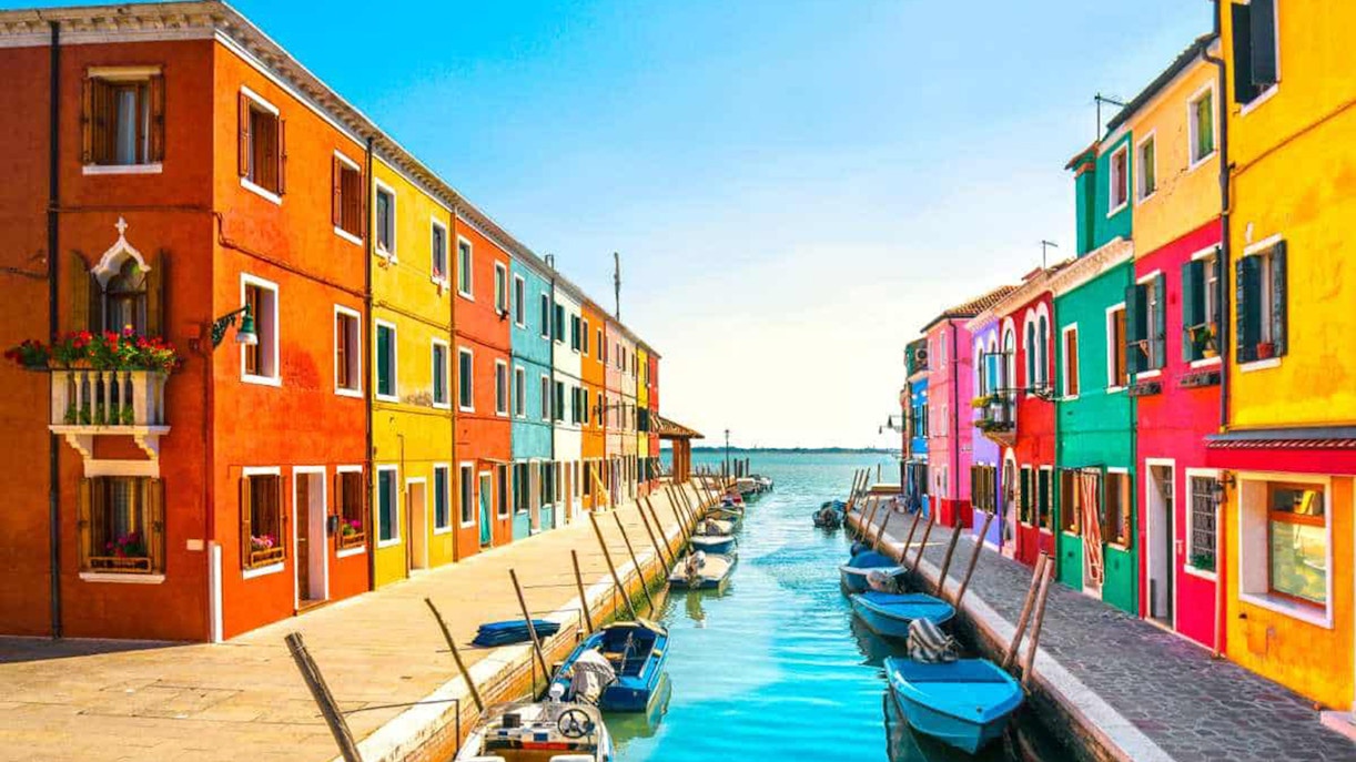 Colorful buildings along a canal in Burano, Venice, with boats moored on the water.