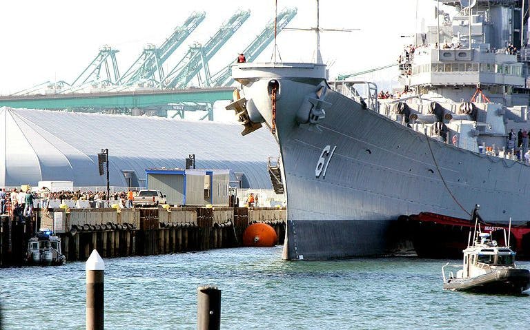 Battleship docked at Los Angeles port with cranes in the background.