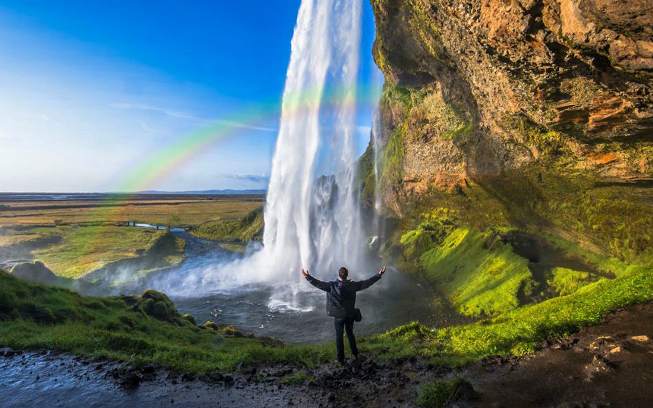 Person standing near Seljalandsfoss waterfall with rainbow, South Coast Classic Day Tour, Iceland.