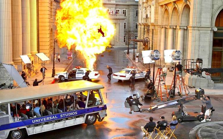 Universal Studios tour tram passing a movie set with explosion and film crew in action.