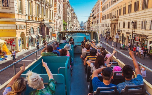 Open-top bus tour on a busy street in Rome, Italy, with tourists enjoying city views.