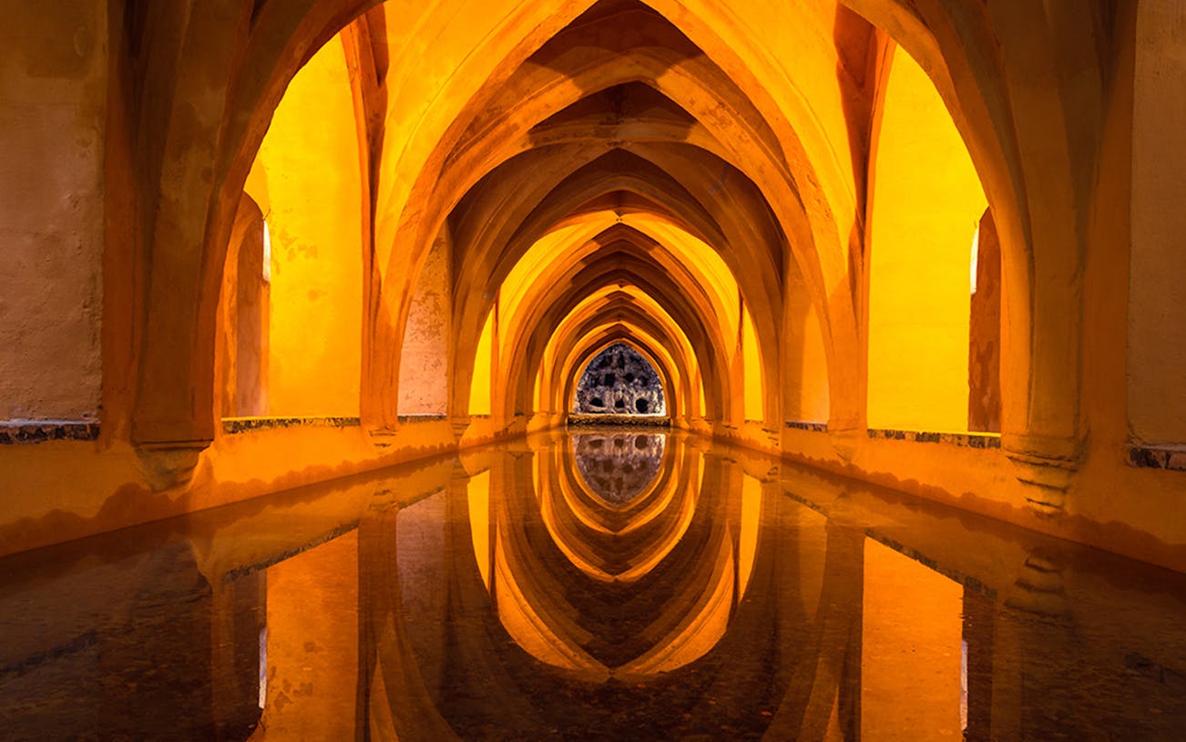 Alcázar of Seville underground baths with arched ceilings and reflections in water.