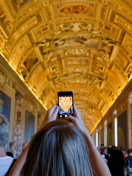 Visitors photographing ornate ceiling in Vatican Museums hallway.