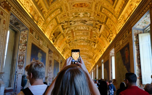 Visitors photographing ornate ceiling in Vatican Museums hallway.