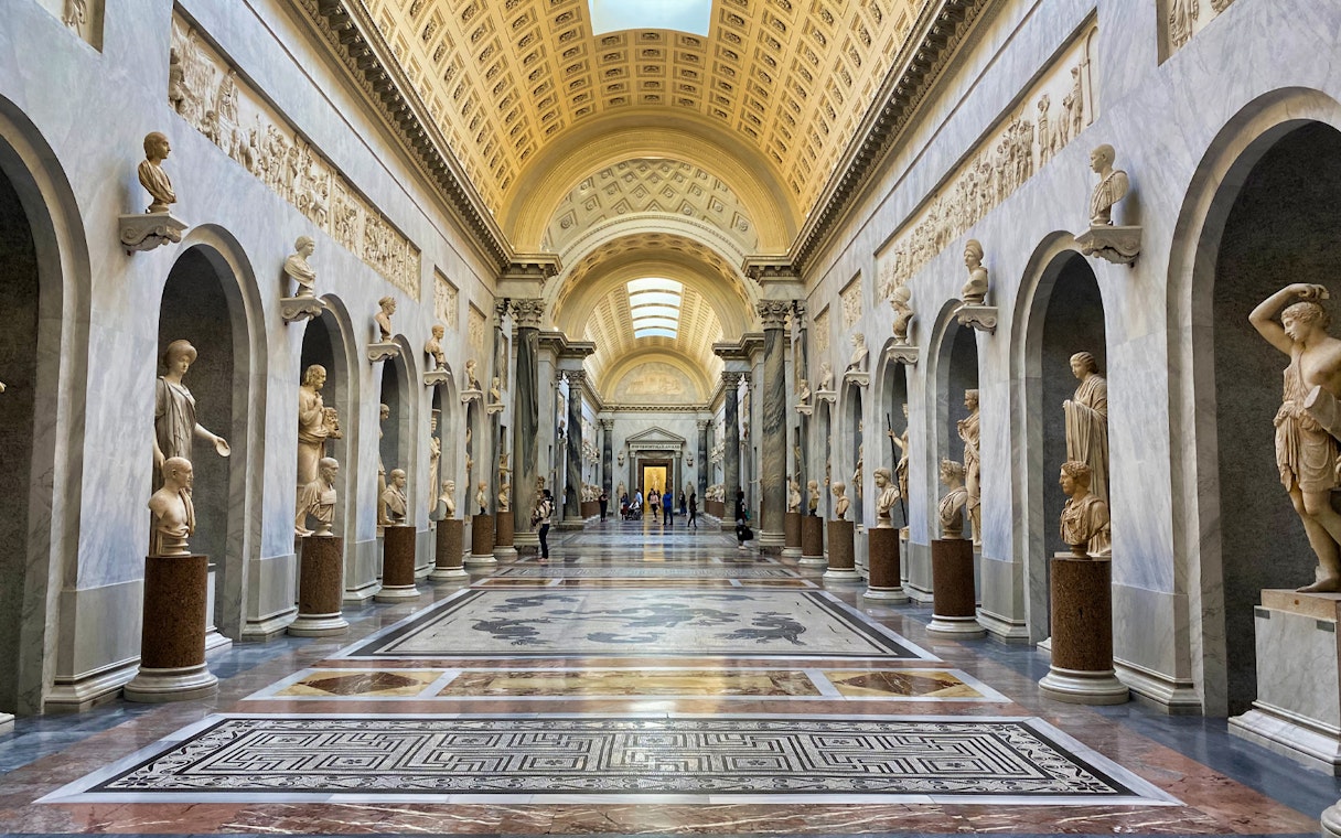 Vatican Museums hallway with classical sculptures and ornate ceiling.