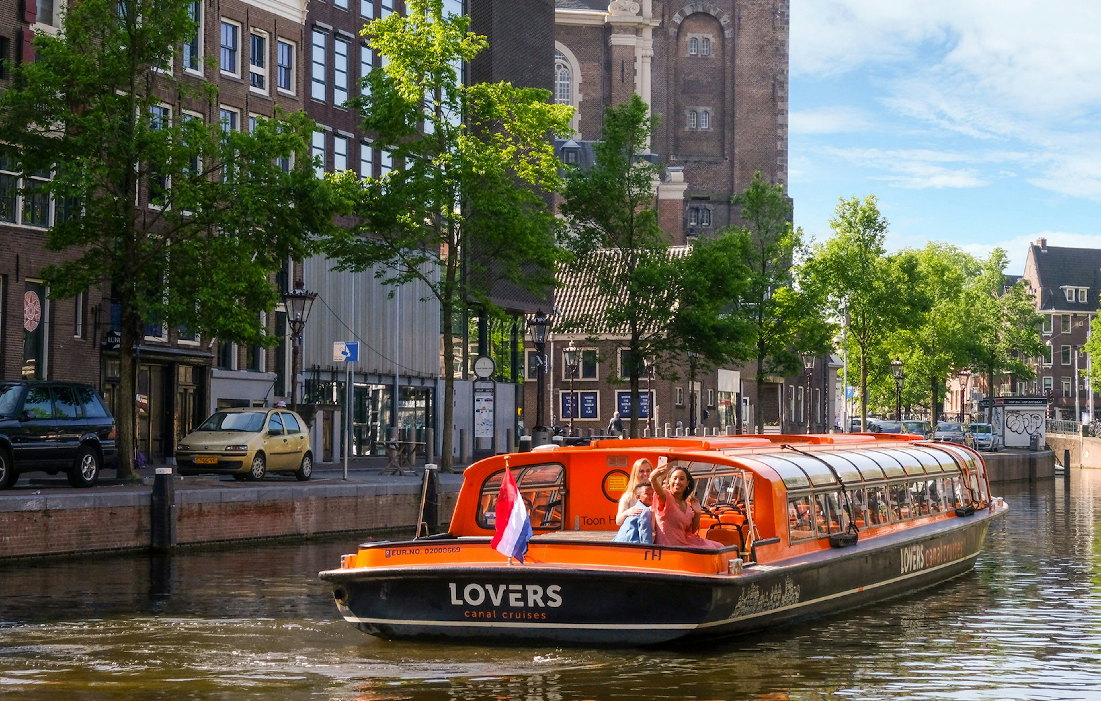 Sightseeing boat on Amsterdam canal with passengers waving, historic buildings in background.