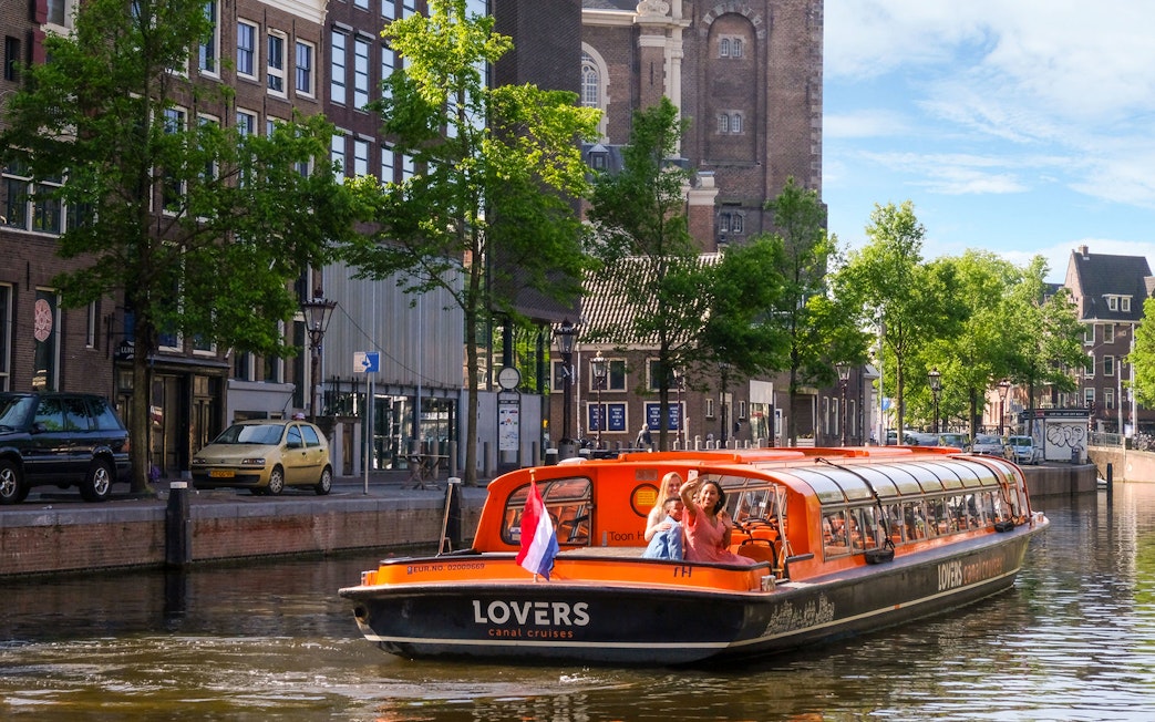 Sightseeing boat on Amsterdam canal with passengers waving, historic buildings in background.