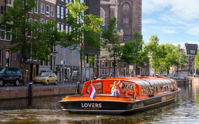 Sightseeing boat on Amsterdam canal with passengers waving, historic buildings in background.