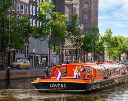 Sightseeing boat on Amsterdam canal with passengers waving, historic buildings in background.