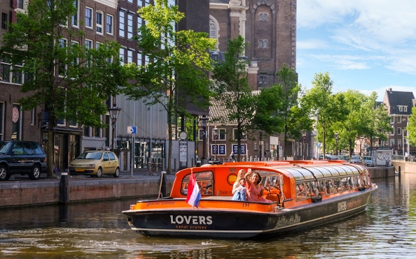 Sightseeing boat on Amsterdam canal with passengers waving, historic buildings in background.