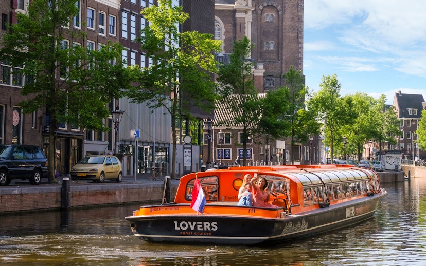 Sightseeing boat on Amsterdam canal with passengers waving, historic buildings in background.
