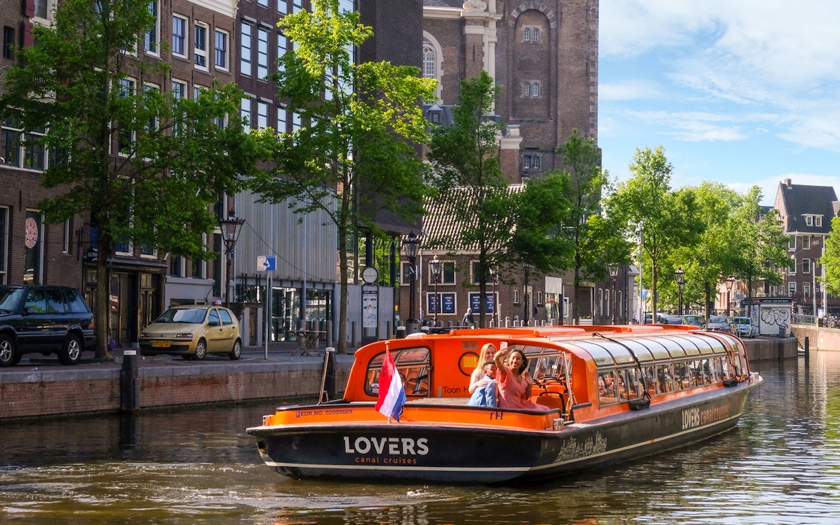 Sightseeing boat on Amsterdam canal with passengers waving, historic buildings in background.