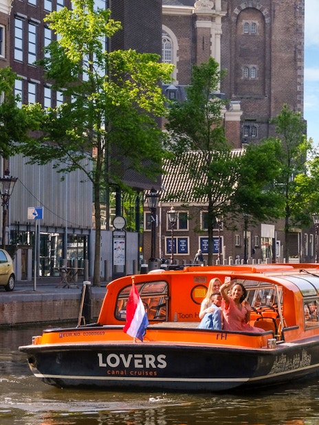Sightseeing boat on Amsterdam canal with passengers waving, historic buildings in background.