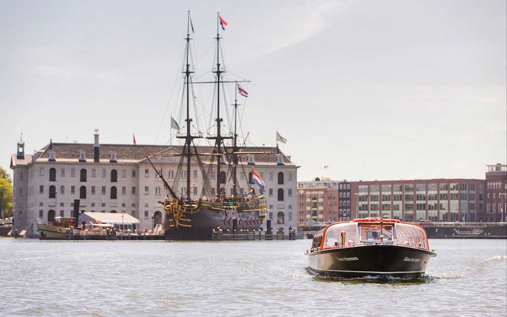 Canal cruise boat on Amsterdam waterway with historic ship and building in background.