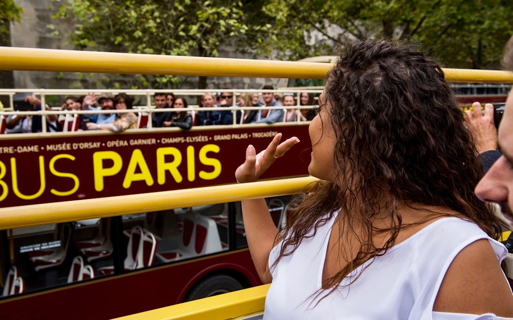 Tourists on Big Bus Paris tour waving, with Eiffel Tower nearby.