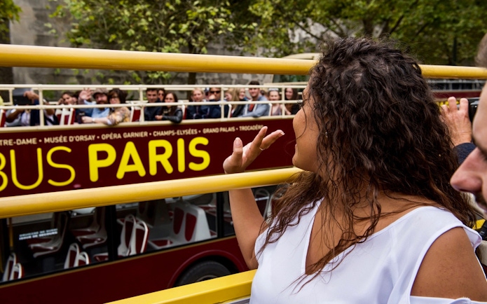 Tourists on Big Bus Paris tour waving, with Eiffel Tower nearby.
