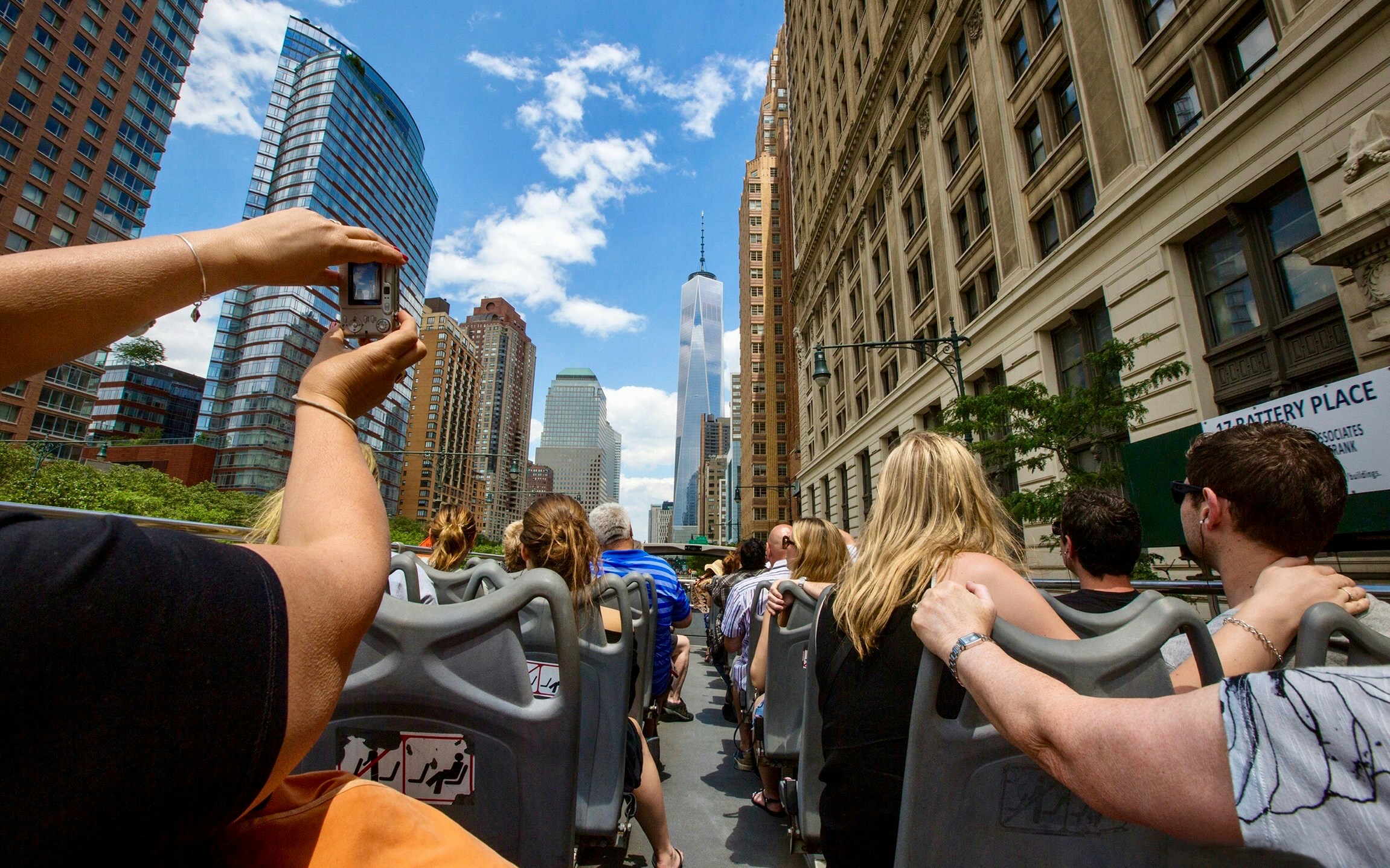 Tourists on a New York City hop-on hop-off bus with Empire State Building and Statue of Liberty in view.