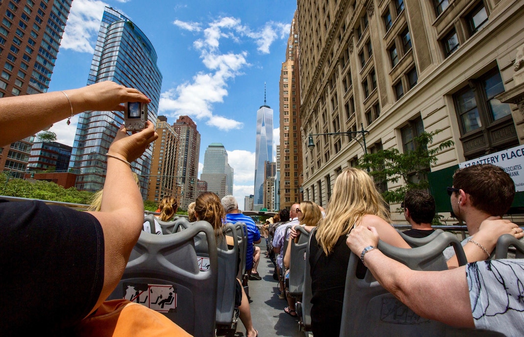 Tourists on a New York City hop-on hop-off bus with Empire State Building and Statue of Liberty in view.