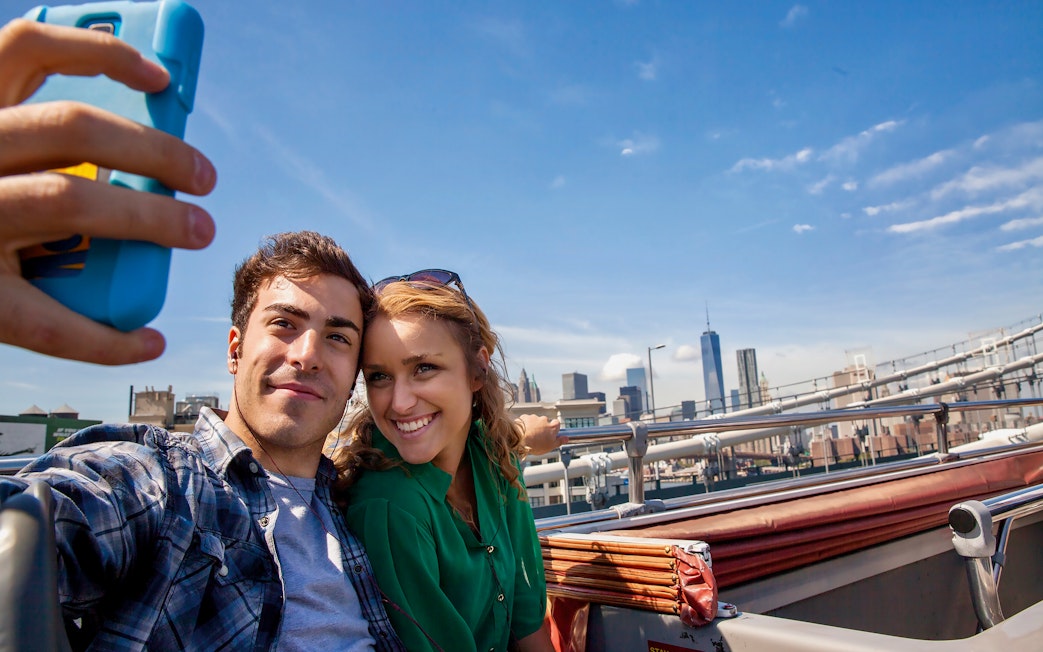 Couple taking selfie on open-top bus with New York skyline in background.