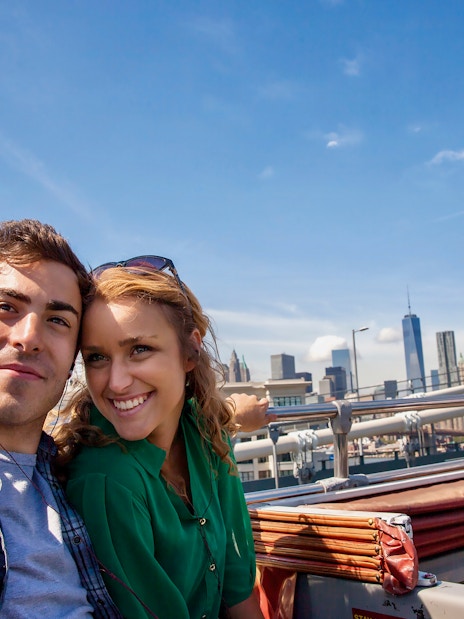 Couple taking selfie on open-top bus with New York skyline in background.