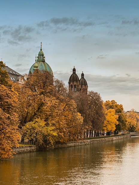 Munich river view with autumn trees and historic buildings, part of Neuschwanstein tour.