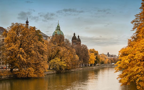 Munich river view with autumn trees and historic buildings, part of Neuschwanstein tour.
