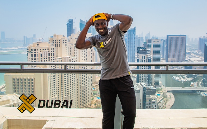 Man smiling on a balcony overlooking Dubai Marina skyline, wearing XLine shirt and cap.