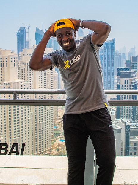 Man smiling on a balcony overlooking Dubai Marina skyline, wearing XLine shirt and cap.