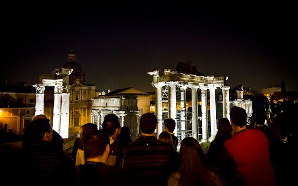 Tour group viewing illuminated Roman Forum ruins at night in Rome.