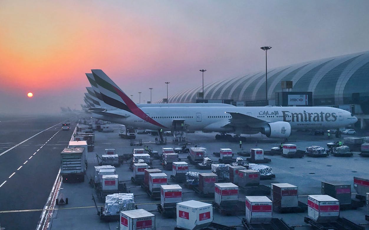 Emirates plane at Dubai Airport Terminal 3 during sunrise with cargo loading.