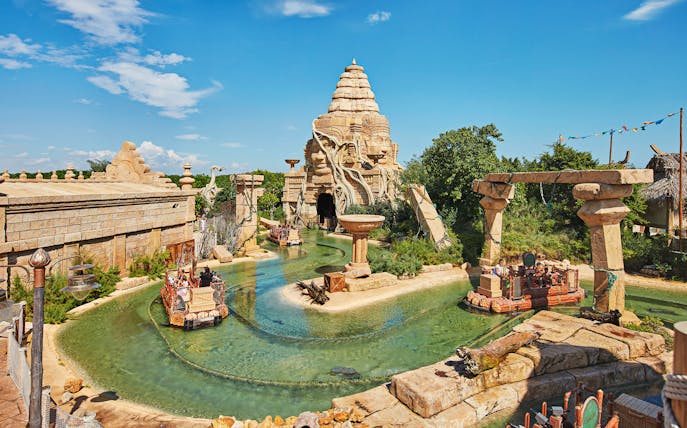 Visitors on a boat ride at PortAventura Park, featuring ancient temple ruins and waterway.
