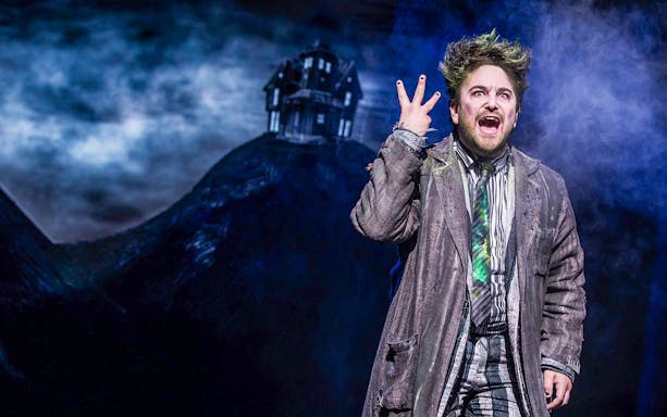 Performer in Beetlejuice costume on stage with eerie house backdrop.
