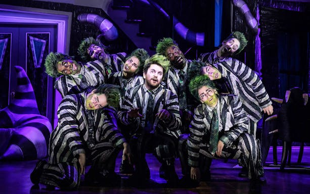 Performers in striped suits and green wigs on stage in a Beetlejuice musical scene.