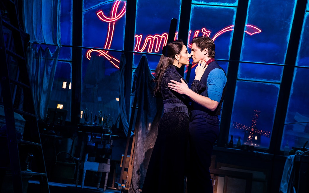Couple embracing on stage with Moulin Rouge sign in background.